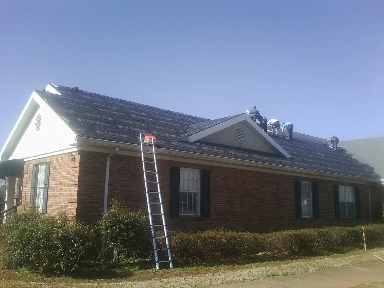 Crew tearing shingles off a brick ranch roof with a ladder set — Emergency Roof Repair work by our crew in Crown Point