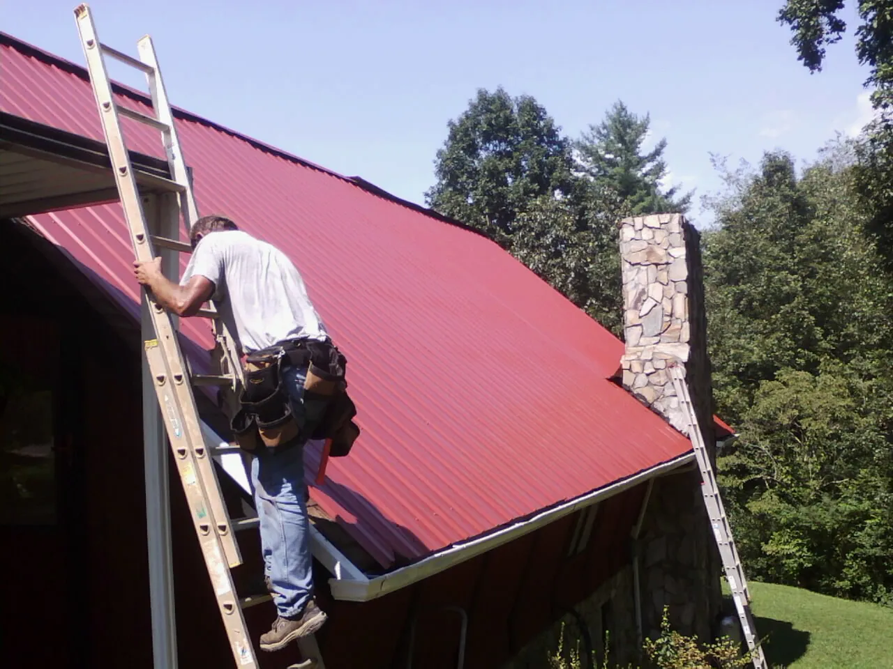Roofer on a ladder beside a red metal roof and stone chimney — Soffit & Fascia Repair by Roofing Brothers in Crown Point